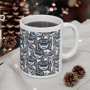 May include: White ceramic coffee mug with a black and blue pattern of stylized faces. The mug is filled with coffee and has a handle on the right side.