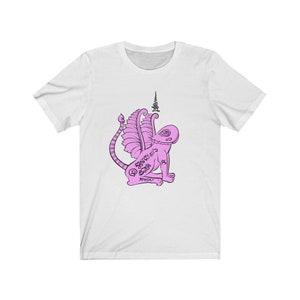 Può includere: Maglietta bianca con un'illustrazione rosa di una creatura mitologica con ali e coda. La creatura è ricoperta di disegni e simboli intricati. Il testo "จตุคามรามเทพ" è scritto sotto la creatura.