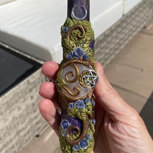 May include: A double-terminated crystal wand featuring a dark purple amethyst point and a white moonstone base. The center is wrapped in green textured clay with bronze swirls, small blue roses, a silver crescent moon, and a silver pentacle charm.