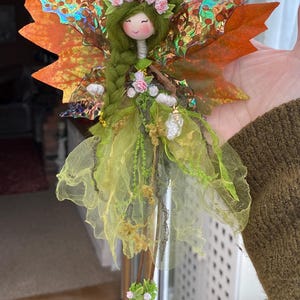 May include: A handmade fairy doll with a peaceful expression, featuring a green braided hairstyle, a floral crown, and delicate wings with orange and iridescent accents. The fairy wears a flowing green and yellow dress, adorned with floral details.