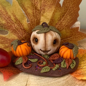 Peut inclure: Une figurine automnale fantaisiste représentant une citrouille blanche avec un visage souriant, entourée de plus petites citrouilles orange et de feuilles brunes, vertes et violettes. La figurine est posée sur une base brune avec des accents verts et violets irisés.
