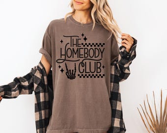 May include: A brown t-shirt with the text "THE HOMEBODY CLUB" in black script and a peace sign hand skeleton graphic. The shirt has a relaxed fit and is worn with a black and tan plaid shirt.