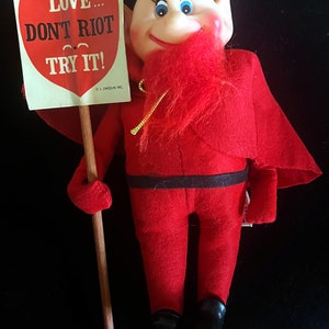 May include: A red devil plush toy holding a sign that says 'LOVE... DON'T RIOT & TRY IT!' The sign is in the shape of a heart. The toy is wearing a red suit with a black belt and black shoes.