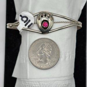 May include: A silver-toned cuff bracelet featuring a bear paw design with a central pink gemstone. The bracelet is displayed on a white surface with a quarter coin for size comparison.