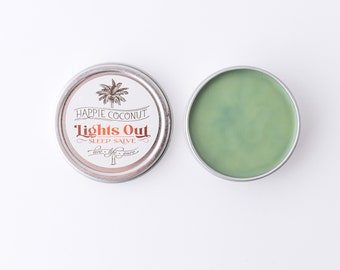 May include: A small, silver tin with a green lid. The tin is labeled "Happie Coconut Lights Out Sleep Salve" with a palm tree graphic. The lid is open, revealing a green salve inside.