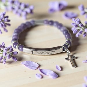 May include: A purple beaded bracelet with a silver cross charm and a silver bar engraved with a cross and the text "I Am With You Always. Matthew 28:20". The bracelet is surrounded by purple flower sprigs and petals.