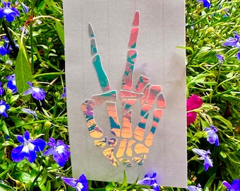 May include: A holographic sticker of a skeleton hand making the peace sign. The sticker has a rainbow iridescent finish.
