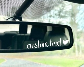 May include: White vinyl sticker with the text "custom text" and a small heart, placed on the rearview mirror of a car.