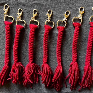 May include: Seven red braided keychains with gold-colored heart-shaped clasps. Each keychain features a braided design that transitions into a tassel at the bottom. The keychains are arranged on a dark gray surface.