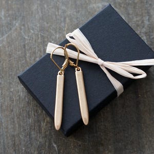 May include: A pair of gold-colored, rectangular drop earrings with lever-back closures. The earrings are displayed on a black gift box tied with a cream-colored ribbon. The background is a textured, gray surface.