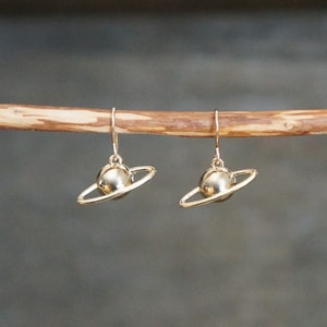 May include: A pair of gold-toned earrings featuring a small sphere with a ring around it, resembling a planet with a ring system.
