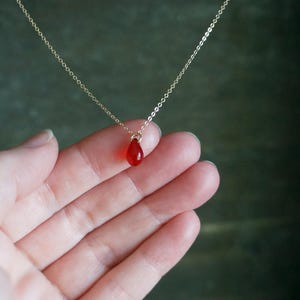 May include: A delicate gold chain necklace featuring a teardrop-shaped, translucent red pendant. The pendant is suspended from a fine chain, creating a simple yet elegant design. The necklace is shown against a neutral background.
