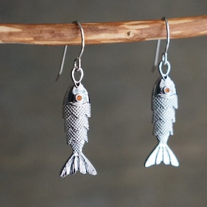 May include: A pair of silver fish-shaped dangle earrings. Each earring features a detailed fish design with textured scales and a small orange gem for the eye. The earrings hang from simple silver hooks.