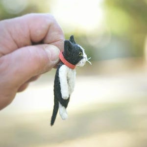 Puede incluir: Figura de gato de fieltro hecha a mano en blanco y negro, con un collar rojo. El gato tiene bigotes blancos y lo sostiene una mano sobre un fondo borroso verde y amarillo. El gato mide aproximadamente 5 cm de alto.