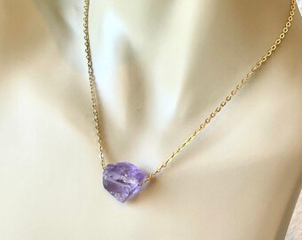 May include: A gold-colored chain necklace featuring a raw, faceted amethyst gemstone pendant. The pendant is a light purple color and is the focal point of the necklace. The chain is delicate and appears to be made of small, linked segments.