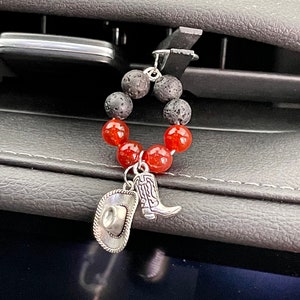 Può includere: Un ciondolo per bocchetta dell'aria dell'auto con un anello di perline rosse e nere. Ciondoli argentati a forma di cappello e stivale da cowboy pendono dall'anello. Il ciondolo è attaccato alla bocchetta dell'aria dell'auto.
