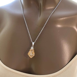 May include: A silver chain necklace with a pendant featuring a light orange, wire-wrapped stone. The stone has a natural, irregular shape. The necklace is displayed on a mannequin.