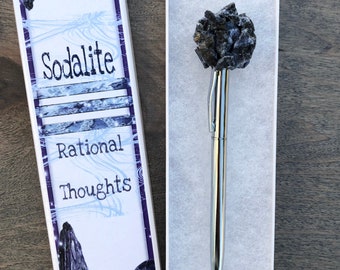 May include: A silver pen topped with a sodalite gemstone, displayed in a white box. The box is decorated with the words "Sodalite" and "Rational Thoughts" and a dark blue and white design. The brand name "justbreathezen" is also visible.