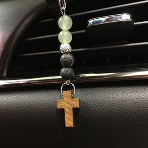 May include: Car air freshener with a wooden cross pendant. The air freshener features a chain with light green beads, a silver beaded spacer, and black lava beads. The cross is made of light brown wood.