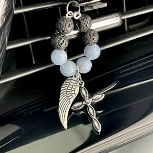 May include: A car air vent diffuser featuring a ring of black lava rock and light blue beads. A silver-colored charm with a wing and a cross hangs from the ring. The diffuser is designed to clip onto a car's air vent.