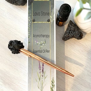 Könnte beinhalten: Ein roségoldener ätherischer Öl-Stift mit einem schwarzen Lavastein-Aufsatz, der auf einer weißen Oberfläche neben einer Schachtel mit der Aufschrift "Lava Stone Aromatherapy Essential Oil Pen" liegt. Die Schachtel zeigt botanische Illustrationen und den Text "JustBreatheZen".