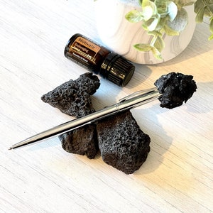 Könnte beinhalten: Ein silberner Stift liegt auf zwei dunklen, porösen Steinen. Eine kleine braune Flasche mit der Aufschrift "Motivate" befindet sich ebenfalls in der Nähe. Der Hintergrund zeigt einen weißen und grauen Pflanztopf mit grünem Laub auf einer weißen Holzoberfläche.