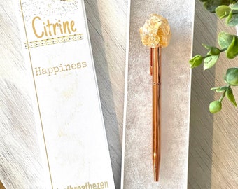 May include: A rose gold pen with a large, yellow citrine crystal top is displayed in a white box. The adjacent box features the words "Citrine" and "Happiness". The box also has the text "Justbreathezzen".
