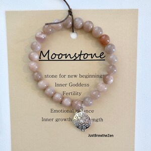 Può includere: Un braccialetto di perline di pietra di luna con un ciondolo d'argento, esposto su una carta beige. La carta recita "Moonstone" ed elenca proprietà come "pietra per nuovi inizi" e "Inner Goddess". Il braccialetto è infilato su un cordoncino marrone.