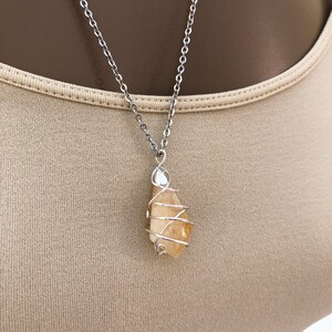 May include: A silver-colored chain necklace with a pendant. The pendant is a light orange-yellow crystal wrapped in silver-colored wire. The necklace is displayed against a neutral-colored background.