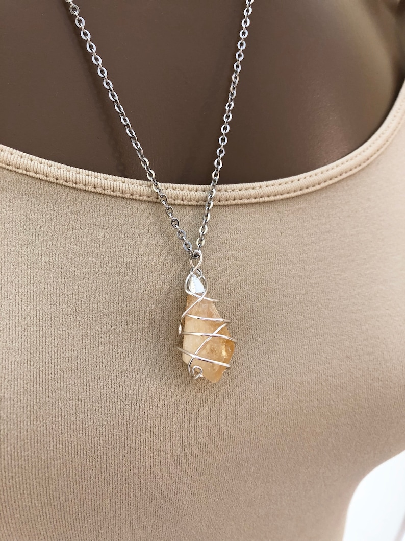 May include: A silver-colored chain necklace with a pendant. The pendant is a light orange-yellow crystal wrapped in silver-colored wire. The necklace is displayed against a neutral-colored background.