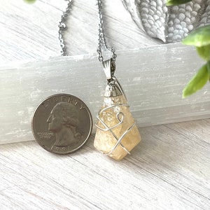 May include: A silver chain necklace with a pale yellow crystal pendant. The crystal is wire-wrapped and has a rough, natural texture. A U.S. quarter coin is next to the pendant for size comparison. The background is a light-colored wooden surface.