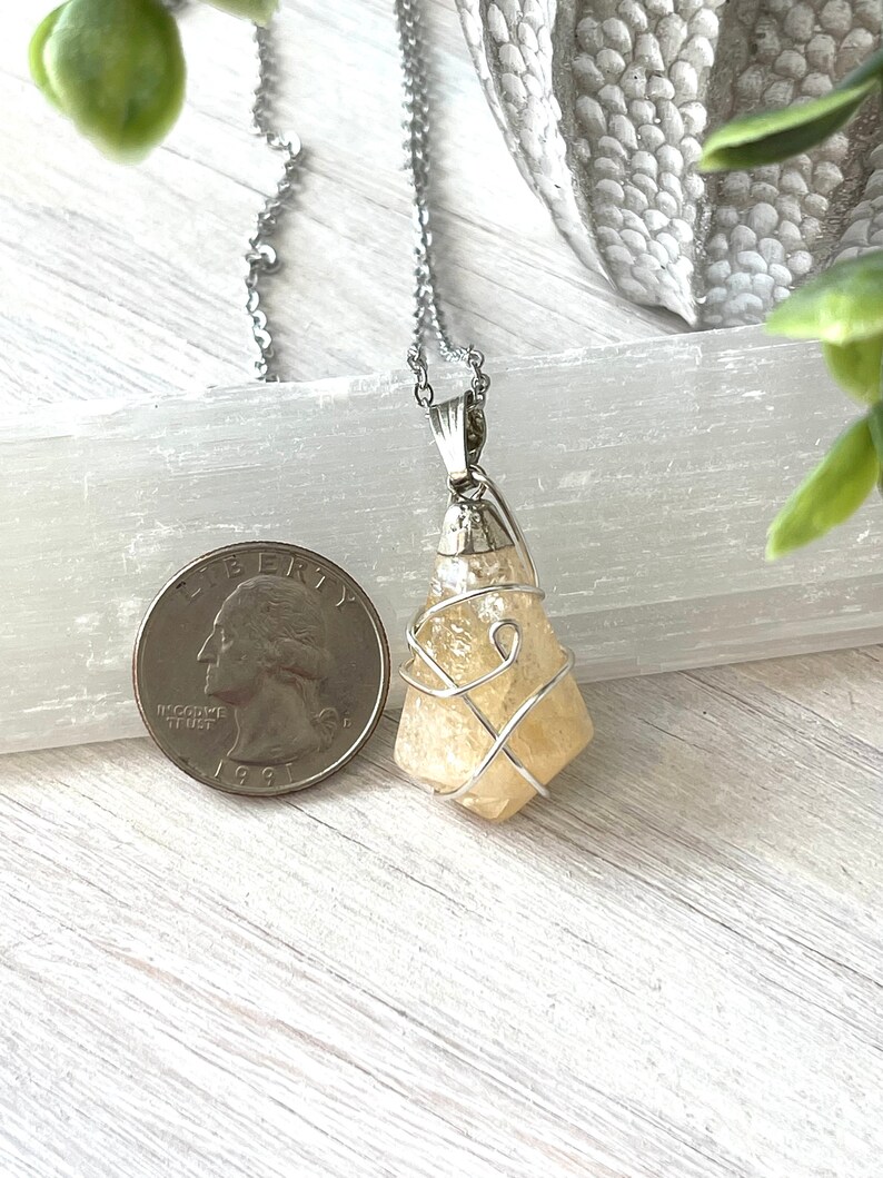May include: A silver chain necklace with a pale yellow crystal pendant. The crystal is wire-wrapped and has a rough, natural texture. A U.S. quarter coin is next to the pendant for size comparison. The background is a light-colored wooden surface.