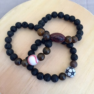 May include: Three beaded bracelets featuring sports-themed charms. One bracelet has a football charm, another a baseball, and the third a soccer ball. The bracelets are made with black lava beads and wooden beads.
