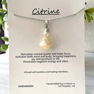 May include: A Citrine pendant necklace with a silver chain. The pendant is a light yellow crystal wrapped in silver wire. The card behind the necklace reads "Citrine" and describes its properties. The card also includes the text "JustBreatheZen" and "Handcrafted by a Mama in Los Angeles, CA."