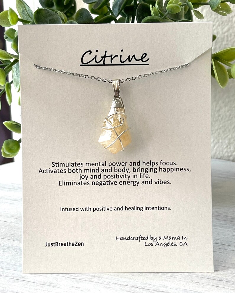 May include: A Citrine pendant necklace with a silver chain. The pendant is a light yellow crystal wrapped in silver wire. The card behind the necklace reads "Citrine" and describes its properties. The card also includes the text "JustBreatheZen" and "Handcrafted by a Mama in Los Angeles, CA."