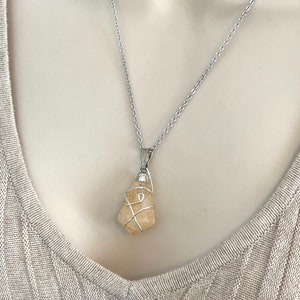 May include: A silver necklace featuring a raw, light yellow citrine crystal pendant. The crystal is wrapped in silver wire and hangs from a delicate silver chain. The necklace is displayed on a neutral-toned background.