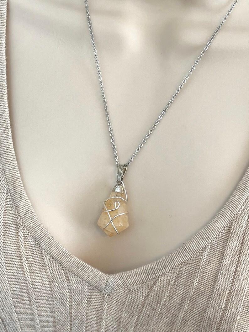 May include: A silver necklace featuring a raw, light yellow citrine crystal pendant. The crystal is wrapped in silver wire and hangs from a delicate silver chain. The necklace is displayed on a neutral-toned background.