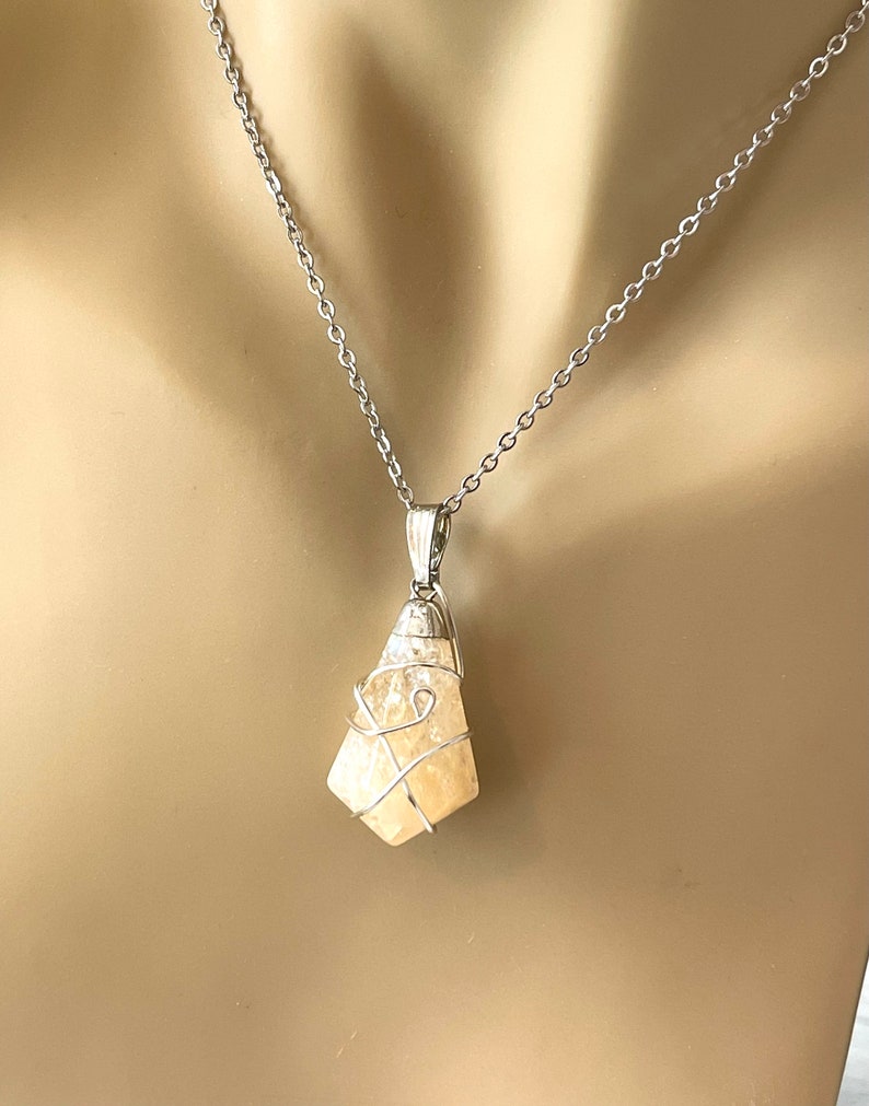 May include: A silver-toned necklace featuring a pendant with a light yellow, irregularly shaped stone. The stone is encased in a silver wire cage and hangs from a delicate chain. The pendant has a polished finish, and the chain is simple and elegant.