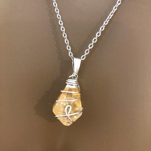 May include: A silver chain necklace with a pendant featuring a raw, yellow-orange citrine crystal wrapped in silver wire. The crystal is teardrop-shaped and hangs from a silver bail.