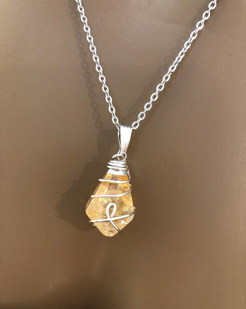May include: A silver chain necklace with a pendant featuring a raw, yellow-orange citrine crystal wrapped in silver wire. The crystal is teardrop-shaped and hangs from a silver bail.