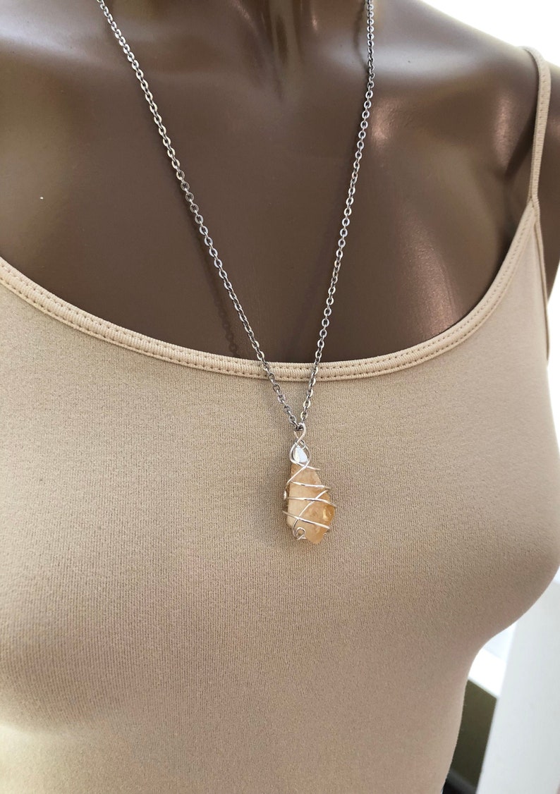 May include: A silver chain necklace with a pendant. The pendant is a light orange stone wrapped in silver wire. The necklace is displayed on a person wearing a beige tank top.