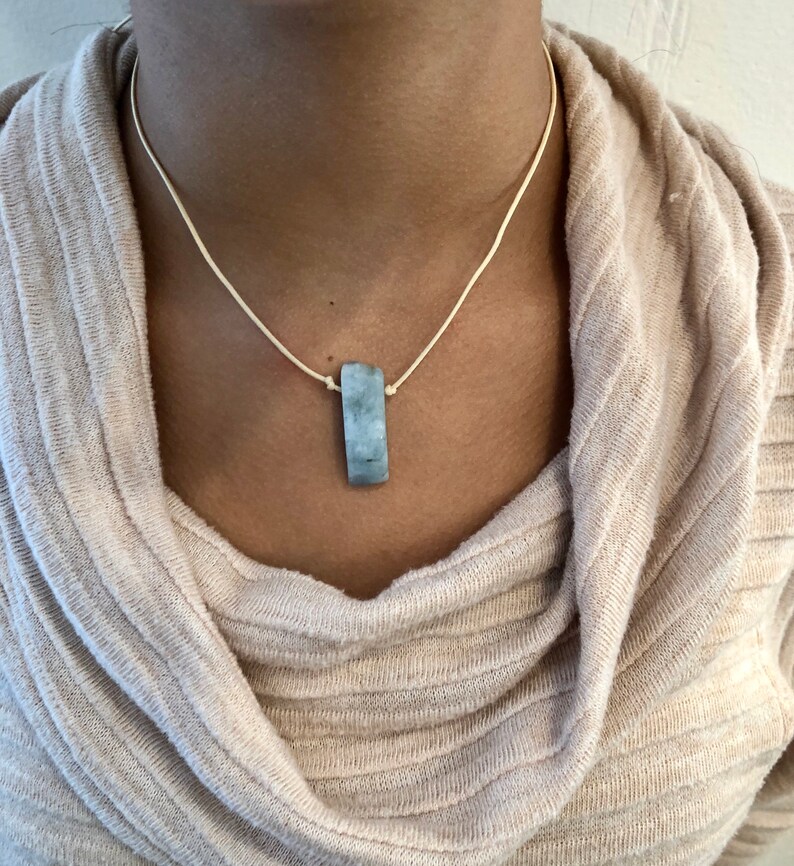 May include: A rectangular pendant necklace with a light blue stone. The stone is suspended from a cream-colored cord. The necklace is worn against a beige sweater.