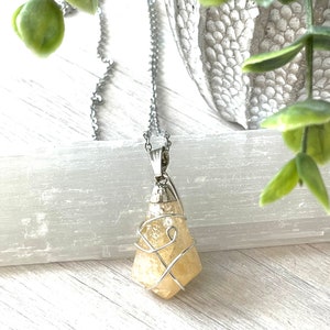 May include: A silver-toned necklace with a pendant featuring a light yellow, irregularly shaped crystal wrapped in silver wire. The pendant hangs from a delicate chain. Green foliage and a white background provide a natural backdrop.