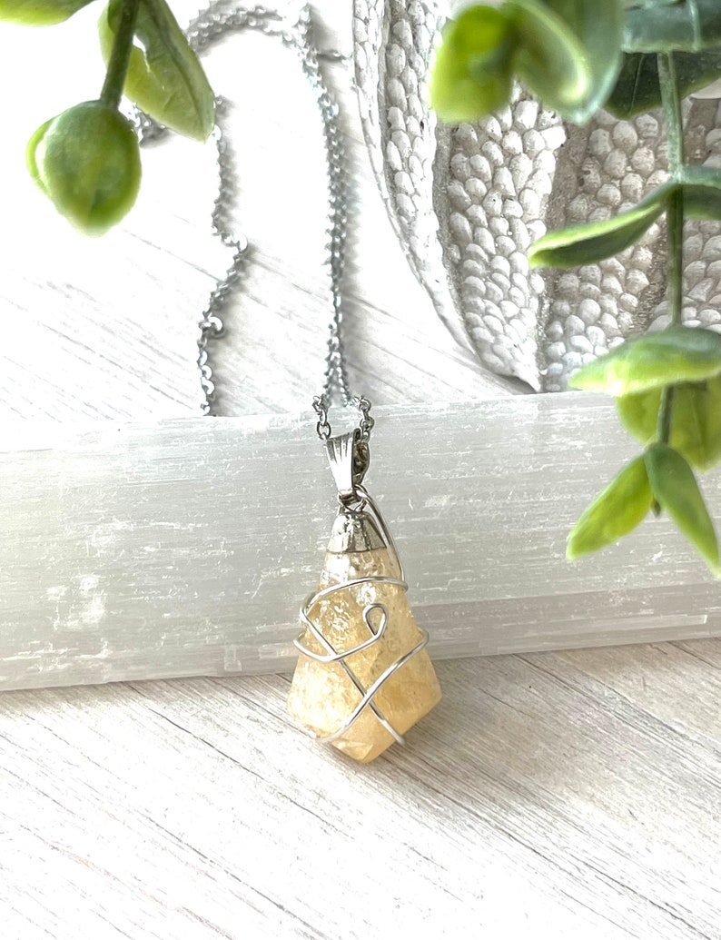 May include: A silver-toned necklace with a pendant featuring a light yellow, irregularly shaped crystal wrapped in silver wire. The pendant hangs from a delicate chain. Green foliage and a white background provide a natural backdrop.
