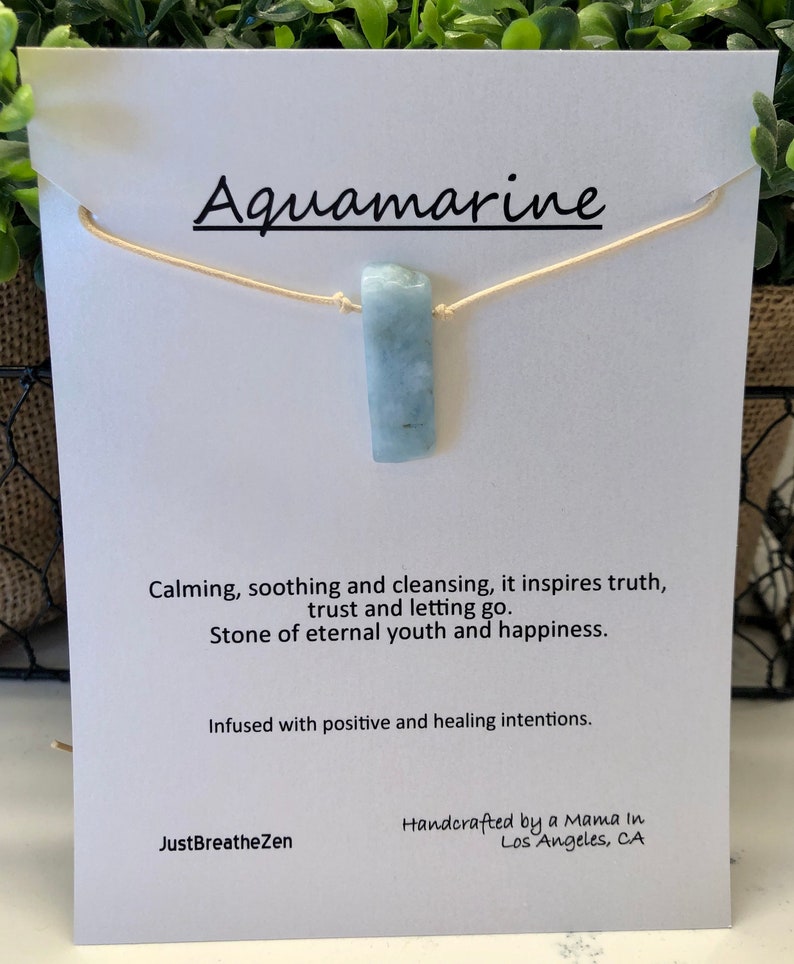 May include: A light blue aquamarine pendant necklace. The rectangular stone is suspended from a beige cord. The card behind the necklace reads "Aquamarine" and includes text about the stone's properties. The card also includes the text "JustBreatheZen".
