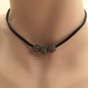 May include: A black choker necklace featuring three gray lava stone beads. The beads have a porous texture and are strung on a thin, black cord. The necklace is displayed on a person's neck.