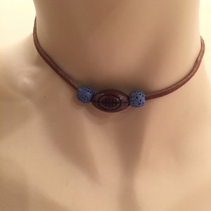 Könnte beinhalten: Eine braune Leder-Choker-Halskette mit einem fußballförmigen Anhänger und blauen Perlenakzenten. Die Halskette wird auf einer Schaufensterpuppe präsentiert, die das Design und die Farbe des Schmucks hervorhebt.