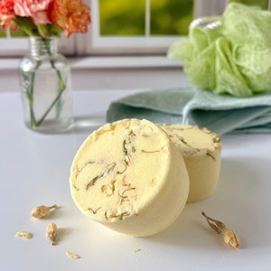 May include: Two round, pale yellow bath bombs with dried flower petals are displayed on a white surface. The bath bombs are decorated with dried petals. A small glass vase with orange and pink flowers is in the background.
