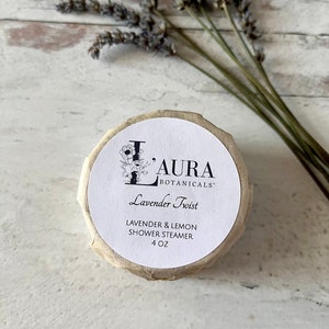 May include: A white round container with a brown paper label. The label says "Laura Botanicals" and "Lavender Twist" in black text. Below the text, it says "Lavender & Lemon Shower Steamer 4 oz".