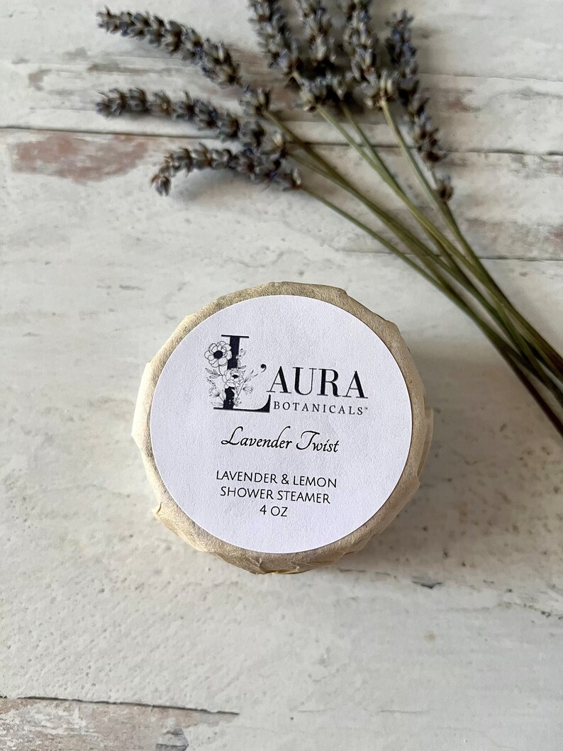 May include: A white round container with a brown paper label. The label says "Laura Botanicals" and "Lavender Twist" in black text. Below the text, it says "Lavender & Lemon Shower Steamer 4 oz".